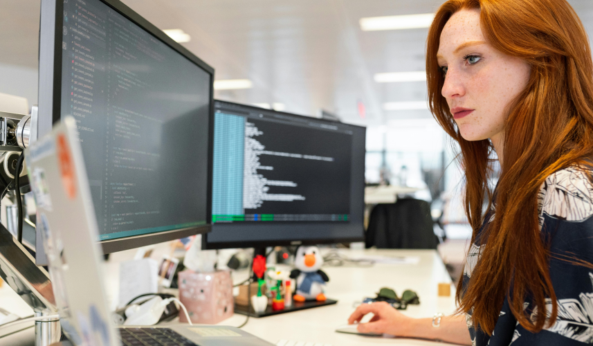 Woman working intently on a computer with dual monitors displaying code.